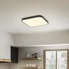 A square LED ceiling light with a black frame and a white diffuser is mounted on a white ceiling in a kitchen setting. Voir les images du produit