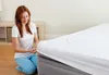 A woman is kneeling on the floor and putting a white sheet on a gray mattress. Produktbild-Vorschau