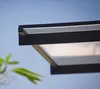 A close-up view of a modern black awning with a white translucent panel, set against a clear blue sky. A blurred green plant is visible in the foreground. Produktbild-Vorschau