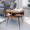 A round wooden side table with black legs, placed next to a gray sofa in a living room setting. The table has a shelf and is decorated with books, headphones, and a vase with sticks. Anteprima dell’immagine prodotto