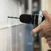 A person's hand is holding a cordless drill and drilling into a wall. Dust is flying from the hole. Produktbild-Vorschau