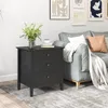 A black nightstand with three drawers sits next to a grey sofa in a living room setting. Anteprima dell’immagine prodotto