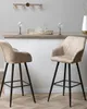 Two beige bar stools are shown in a modern setting, with a bar counter and decorative items in the background. Náhled obrázku produktu