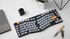 A split ergonomic keyboard with gray and orange keycaps is shown on a white desk, next to a notebook, an inkwell, and a fountain pen. Produktbild-Vorschau