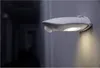 A white LED outdoor light fixture with a motion sensor is mounted on a gray wall. The light is illuminated, casting a warm glow downwards. The background is dark and out of focus, suggesting a hallway or exterior setting at night. Voir les images du produit