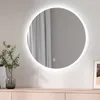 A round LED mirror with a white light ring is mounted on a light gray wall above a light pink dresser. A vase with green leaves is on the left side of the dresser. Produktbild-Vorschau