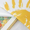 A close-up view of a fabric with a yellow abstract design, partially covering a framed artwork. Text is visible on a separate piece of paper in the foreground. Produktbild-Vorschau