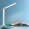 A white LED desk lamp is shown on a light blue background, with a book and a smartphone charging wirelessly in the foreground. Produktbild-Vorschau