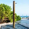 An outdoor shower on a beach next to lush green foliage, with lounge chairs and umbrellas visible in the background by the ocean. Produktbild-Vorschau