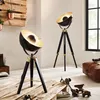 Two industrial-style tripod floor lamps are shown in a room with a wooden pallet wall and a leather ottoman. Podgląd zdjęcia produktu