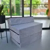 A gray folding mattress is shown in a room with large windows overlooking a balcony with greenery. Náhled obrázku produktu