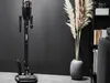 A cordless vacuum cleaner is displayed on its charging stand next to a vase with dried pampas grass and a bookshelf. Produktbild-Vorschau