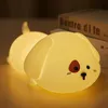 A cute, illuminated puppy-shaped night light is lying on a surface, with books in the background. Produktbild-Vorschau