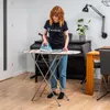 A woman is ironing clothes on an ironing board in a room with a piano and dining table. Produktbild-Vorschau