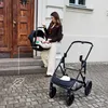 A woman is holding a baby car seat next to a stroller, standing on a cobblestone path in front of a building with a wooden door. Produktbild-Vorschau