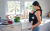 A woman is holding a baby in a carrier in a kitchen. Produktbild-Vorschau