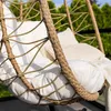 Close-up of a hanging chair with white cushions and a beige woven exterior, set against a blurred background of green foliage and a light sky. Produktbild-Vorschau