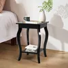 A black wooden nightstand with two shelves is placed next to a bed. A mug, keys, and books are on the top shelf, and books are on the bottom shelf. A vase with greenery is also on the top shelf. Podgląd zdjęcia produktu