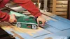 A person is using a Bosch belt sander to remove paint from a wooden surface. Produktbild-Vorschau