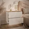 A modern white high-gloss nightstand with gold trim and LED lighting, placed next to a bed in a bedroom setting. Produktbild-Vorschau