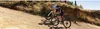Two cyclists riding on a dirt road in a dry, hilly landscape. Produktbild-Vorschau