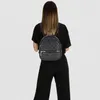 A woman wearing a black outfit is shown from the back, wearing a grey backpack with a repeating logo pattern. Produktbild-Vorschau