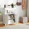 A white wooden cabinet with doors and shelves, decorated with books, a vase of flowers, and a radio. A brown woven bag and an orange cardigan hang on the wall behind it, and a beige ottoman sits to the side. Náhled obrázku produktu