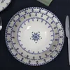 A stack of white ceramic plates and bowls with a blue decorative pattern, laid out on a dark blue tablecloth. Produktbild-Vorschau
