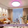 A hand holding a remote control points towards a modern ceiling light in a living room. Produktbild-Vorschau