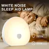 A baby sleeping peacefully next to a white noise sleep aid lamp. Produktbild-Vorschau