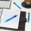 A close-up shot of a blue and black ballpoint pen resting on a notebook, with a laptop and a coffee cup in the background. Podgląd zdjęcia produktu