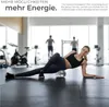A woman is using a foam roller in a gym setting with other people exercising in the background. Produktbild-Vorschau