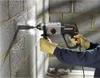 A person wearing yellow gloves and a blue shirt is using a WORX WX339 Bohrhammer to drill into a concrete block wall. Dust and debris are visible around the drill bit. Náhled obrázku produktu