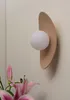 A wall lamp with a beige curved shade and a white spherical light bulb is mounted on a light beige wall. Pink lilies are in the foreground. Podgląd zdjęcia produktu