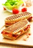 Two panini sandwiches are displayed on a wooden board, with a salad and tomatoes in the background. Podgląd zdjęcia produktu