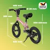 A pink balance bike with various dimensions indicated by lines and text overlays, set against a gradient green and yellow background. A quality seal is visible in the top right corner. Produktbild-Vorschau