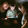 A mother and two children are reading a book in the dark, illuminated by a headlamp and a flashlight. Produktbild-Vorschau