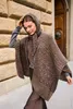 A woman wearing a knitted poncho with sequins, standing outdoors next to a brick wall and a wooden door. Produktbild-Vorschau