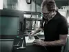 A man is working on a workbench with tools and a metal object. Produktbild-Vorschau