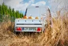 A white trailer filled with pumpkins is parked in a field of dry grass, with a line of green trees in the background. Produktbild-Vorschau