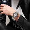 A person wearing a black leather jacket and a white t-shirt, showing off a silver chronograph watch on their wrist. Podgląd zdjęcia produktu