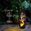 A solar-powered garden light with a tree silhouette is placed on a brick patio, surrounded by lush greenery and an old stone urn. Produktbild-Vorschau