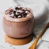 A close-up of a glass mug filled with hot chocolate, topped with marshmallows and chocolate shavings, resting on a cork coaster. A wooden spoon is placed next to the mug. Produktbild-Vorschau