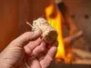 A hand holding a fire starter log in front of a fireplace with flames. Produktbild-Vorschau
