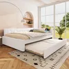 A white daybed with a pull-out trundle bed is set up in a bright, airy room with large windows overlooking a green landscape. The room has a modern, minimalist aesthetic with a textured wall, wooden floor, and a patterned rug. Podgląd zdjęcia produktu