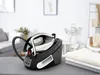 A steam iron station is placed on an ironing board in a bright, modern room. Folded clothes are stacked to the right of the iron. Produktbild-Vorschau