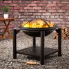 A fire pit with flames burning sits on a gravel surface in front of a brick wall. A small wooden table with a potted plant is to the left, and a stack of firewood is to the right. Produktbild-Vorschau