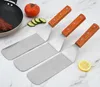 Three spatulas with wooden handles are laid out on a marble-like surface, with a frying pan, steak, tomato, cucumber slices, and a lemon slice visible in the background. Anteprima dell’immagine prodotto