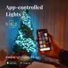 A hand holding a smartphone displaying a light control app, with a Christmas tree decorated with blue lights in the background. Náhled obrázku produktu