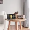 A pet urn with a photo frame and a decorative tree is placed on a round wooden table next to a basket of pinecones and a small radio. A sofa is visible in the background. Náhled obrázku produktu